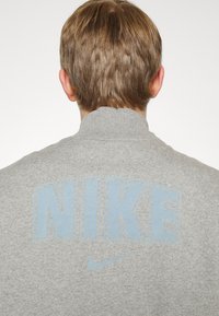 Person med kort brunt hår som bär en grå Nike-sweatshirt med stor bl å "NIKE"-text och swoosh-logotyp på baksidan.