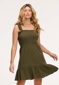 Vestido verde oliva texturizado con escote cuadrado y tirantes con volantes. Bajo acampanado que añade volumen, confeccionado con una tela ligera.