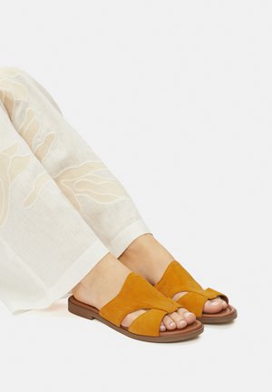 Pomarańczowe zamszowe sandały z krzyżowym designem, odkryty palec i płaska podeszwa, zestawione z jasnymi, wzorzystymi spodniami o szerokich nogawkach.