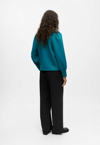 Top lungo a maniche con texture color turchese e spalle arricciate; abbinato a pantaloni neri a vita alta e ampi, con calzature nere.