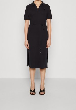 Mujer con vestido midi negro de manga corta con aberturas laterales, cuello, cinturón de lazo y sandalias negras de tacón con punta abierta.