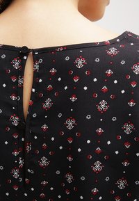 Černá látková blůza s květinovým vzorem v červené a bílé barvě, se zapínáním na knoflík na zádech a klíčovou dírkou.