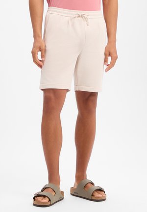 Mężczyzna ubrany w beżowe szorty z troczkami i szare sandały na dwóch paskach, stojący na białym tle.