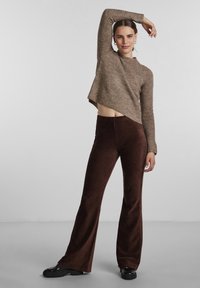 Brown textured cropped sweater with long sleeves paired with brown corduroy flared pants. Model stands with one arm raised and a neutral background.