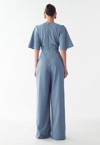 Modra kombinezona z širokimi rokavi, oprijetim telovnikom in ohlapnimi širokimi hlačami. Gladka tekstura, zadrga na hrbtu in brez vidnih vzorcev.