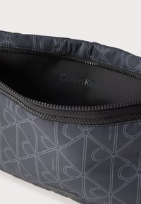 Czarna saszetka Calvin Klein z szarą geometryczną wzorem serca, częściowo rozpięta, ukazująca nazwę marki wewnątrz saszetki.
