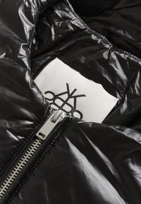 Interior de chaqueta acolchada negra con textura brillante; cuenta con cremallera plateada y etiqueta con diseño gráfico negro sobre fondo blanco.