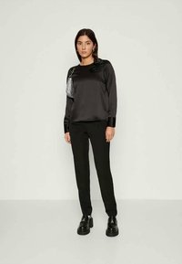 Woman wearing black satin long-sleeve top with flower detail, black pants, platform shoes, and small silver handbag on shoulder.