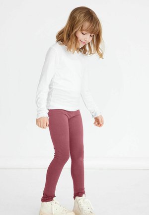 Weißes Langarmoberteil kombiniert mit kastanienbraunen Leggings, die ein figurbetontes Design und eine glatte Textur aufweisen. Das Model trägt weiße Sneakers.