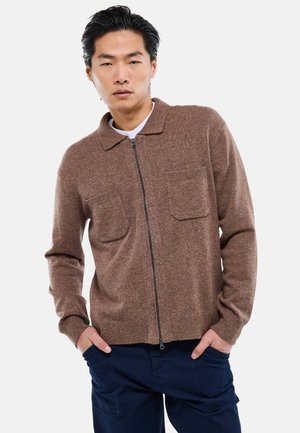 Jeune homme aux cheveux noirs portant un pull zippé marron avec poches et un pantalon bleu marine, mains dans les poches, fond neutre.