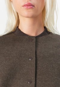 Vaaleahiuksinen nainen, jolla on päällään ruskea napillinen villatakki, jossa on ribbineulekaulus, kuvattuna kaulasta ylävartaloon.