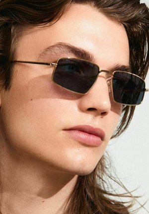 Lunettes de soleil noires rectangulaires avec accents en métal doré, dotées d'une monture fine et de verres teintés, sur un fond clair.