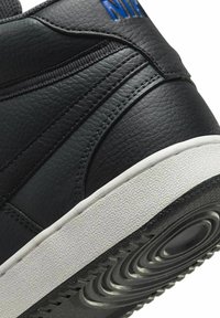 Zapatilla negra de cuero con superficie texturizada, suela de goma blanca y detalle de logo azul. Presenta un diseño de caña alta con acentos de costura.