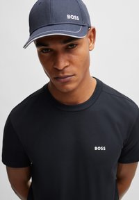 Marinblå keps med "BOSS"-logotyp, i kombination med en svart t-shirt med kort ärm som har en liten vit "BOSS"-logotyp på bröstet.