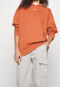 Oranžová oversize mikina s dlouhými rukávy a bílým logem Nike. Párována se světle šedými cargo kalhotami se zipovým kapsou a zlatým logem Nike.