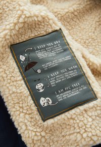 Beige fleecestof heeft een blauw label met witte tekst en illustraties, die de waterdichte eigenschappen en het behoud van warmte beschrijven.