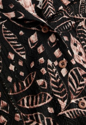 Sort stof med et struktureret mønster af pink blade og geometriske former. Har brune knapper og en krave detalje.