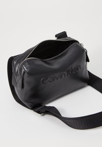 Črna usnjena torbica preko telesa z zadrgo, ki ima odtisnjen logotip "Calvin Klein", nastavljiv trak in gladko teksturo.