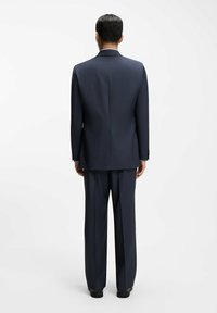 Uomo in piedi di spalle, indossa un completo blu scuro con pantaloni a gamba dritta e scarpe eleganti nere su uno sfondo bianco uniforme.