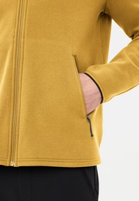 Giacca in pile gialla con zip frontale e due tasche laterali. Presenta una texture liscia, una zip nera a contrasto e una vestibilità sagomata.