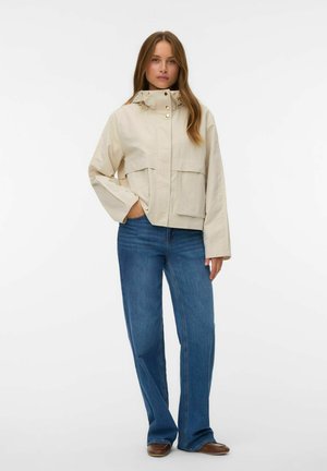 Kvinde stående iført en beige hættetrøjejakke med store lommer, blå bukser med lige ben og brune sko mod en ensfarvet baggrund.