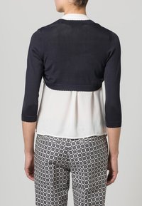 Marinblå kort kavaj med trekvartsärmar, lagd över en vit blus, i kombination med mönstrade byxor i nyanser av blått och krämfärg.