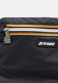 Primo piano di una borsa in tessuto blu navy con una cerniera bianca, uno sfondo a strisce giallo-arancio e un tirante in metallo con il logo K-Way e una patch con il logo.