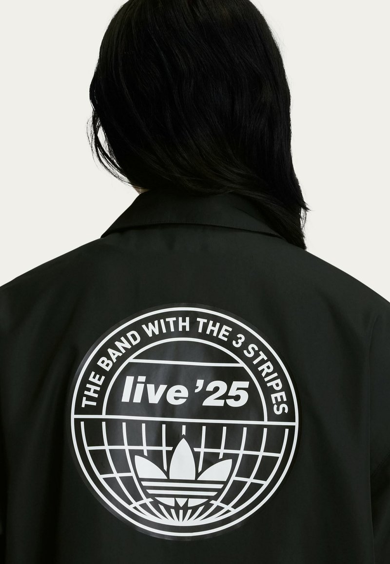 Black jacket featuring a circular white graphic logo with text "THE BAND WITH THE 3 STRIPES" and "live '25" on the back, smooth texture.