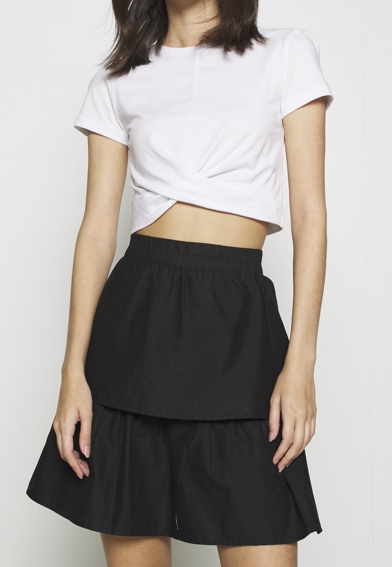 Kvinna klädd i en vit kortärmad croptop och en svart lager-på-lager kjol, stående mot en enfärgad vit bakgrund.