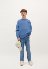 Felpa blu con tasca frontale, abbinata a jeans di denim azzurri. Tiene un cappellino crema con testo scuro e indossa sneakers beige.
