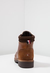 Hnědá kožená kotníková bota s měkkým hnědým límcem, tmavě hnědou podrážkou a červeným, bílým a modrým akcentem na patě. Hladké a strukturované povrchy.