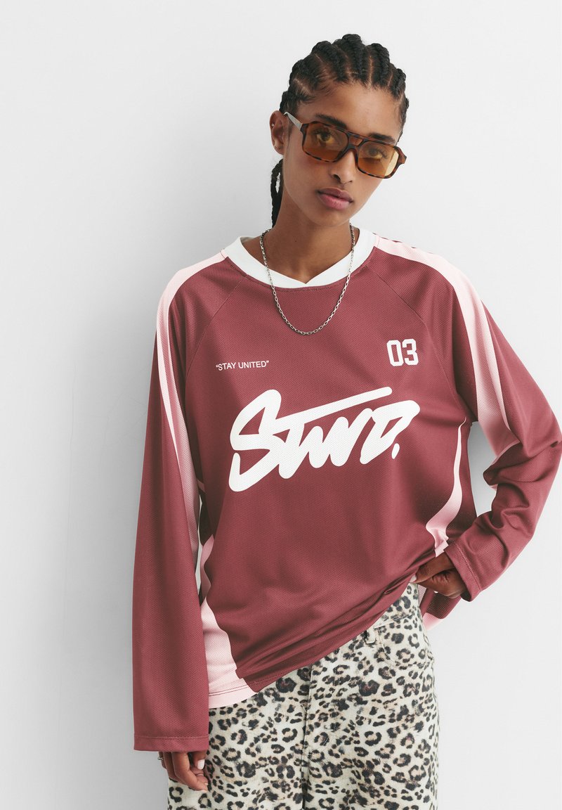 Bordo majica s dolgimi rokavi z belim napisom "Stay United" in logotipom, rožnate akcenti. Hlače v leopardjem vzorcu. Zatemnjena sončna očala in verižica.