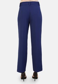 Pantalon ajusté bleu marine avec une coupe droite, des plis discrets et des passants de ceinture. Tissu lisse, porté avec des chaussures à talons.