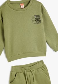 Olivegrön sweatshirt och matchande byxor, tillverkade av mjukt tyg. Sweatshirten har ett svart grafiskt tryck samt ribbade manschetter och nederkant.