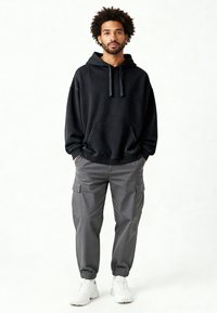 Felpa nera con cappuccio e tasca frontale, abbinata a pantaloni cargo grigi con tasche laterali e polsini elastici, indossati con scarpe da ginnastica bianche.