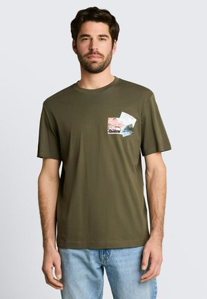 Mann trägt olivgrünes T-Shirt mit kurzen Ärmeln und reisethematischen Drucken auf der linken Brust, kombiniert mit hellblauen Jeans, steht vor einfarbigem Hintergrund.
