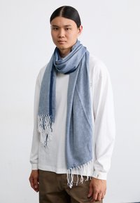 Light blue scarf with a textured pattern, fringed ends, and a dark blue accent band. Worn casually over a white long-sleeve shirt.