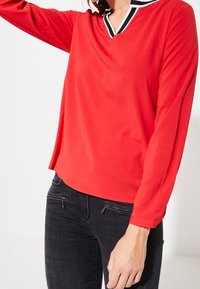 Rød bluse med lange ærmer og V-hals, med en sort og hvid stribet krave. Lavet af glat stof, har den en afslappet pasform.