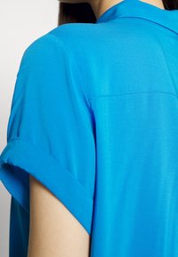 Gros plan sur une personne portant une chemise à manches courtes bleu vif avec les poignets retroussés, montrant le haut du dos et la zone des épaules.