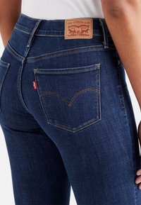 Dark blue denim jeans with a mid-rise fit, featuring a leather label, red stitching, and distinct back pocket design.
