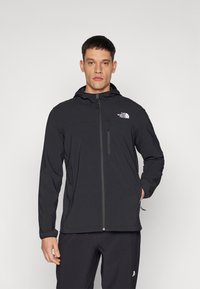 Schwarze Softshell-Jacke mit Kapuze, Frontreißverschluss und seitlichen Taschen. Mit einem Logo auf der Brust und eng anliegenden Bündchen. Strapazierfähige, glatte Textur.