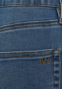 Jeans din denim albastru cu o textură fină, având cusături deschise și un mic logo brodat "IVY" pe buzunarul din spate.
