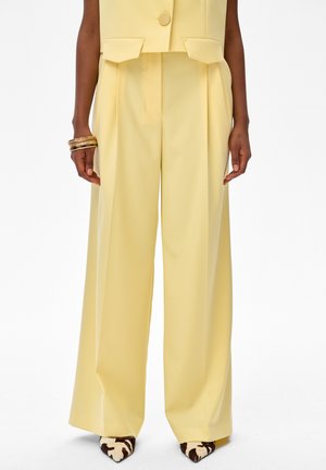 Żółte spodnie z wysokim stanem i szerokimi nogawkami, zestawione z pasującą kamizelką, noszone z czarno-białymi butami z ostrym noskiem i złotymi bransoletkami na jednym nadgarstku.