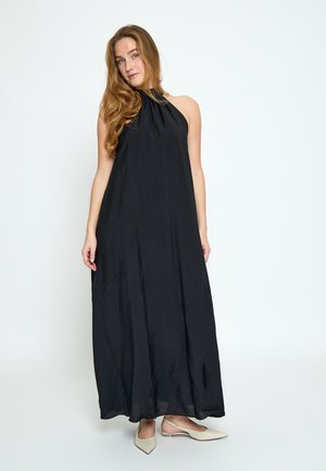 Kobieta z długimi włosami ubrana w czarną maxi suknię na ramiączkach z odsłoniętymi ramionami oraz beżowe, spiczaste baleriny, stojąca na tle jednolitego białego tła.