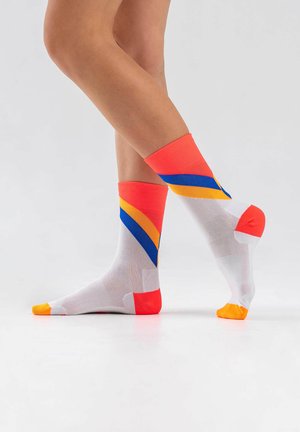 Legs crossed wearing white ankle socks with orange, blue, and yellow diagonal stripes and orange toe and heel accents on a white floor.