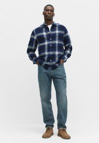 Karierte Flanellhemd in Marineblau mit Knopfkragen und langen Ärmeln. Kombiniert mit hellblauen Jeans und hellbraunen Wildlederschuhen.