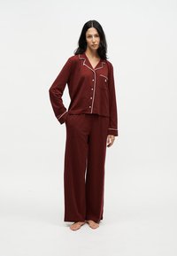 Femme debout pieds nus portant un haut de pyjama bordeaux à manches longues avec boutons et bordure blanche, ainsi qu’un pantalon de pyjama ample assorti.