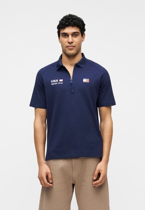 Uomo che indossa una polo blu navy a maniche corte con zip e logo USA Sail GP Team e pantaloni beige, in piedi davanti a uno sfondo bianco.