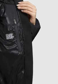 Svart puffjacka med en blank yta, utrustad med en ficka med dragkedja, en logotyp patch och kontrasterande svarta tygdetaljer. Slät textur.