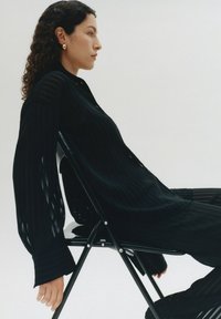 Schwarz gestreiftes Shirt mit langen, locker sitzenden Ärmeln, getragen von einem Model, das auf einem Klappstuhl vor einem schlichten Hintergrund sitzt.